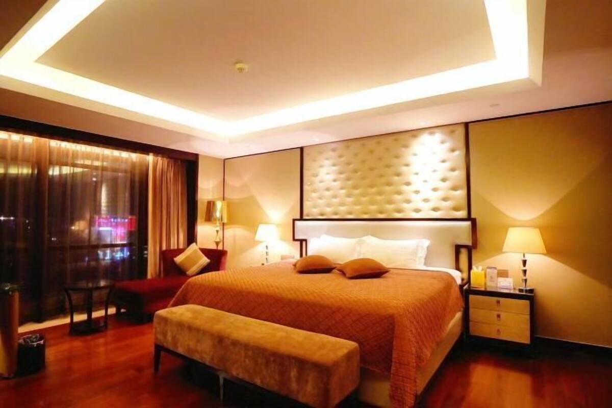 A modern hotel room with a large bed, a bench, a nightstand, and a view of the city at night.