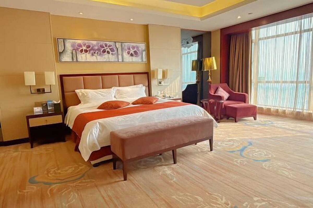 A hotel room with a large bed, a bench, a desk, a chair, and a large window.