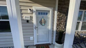 Une maison portant le numéro 201, une porte blanche avec une pancarte « Bienvenue », un paillasson orné de motifs de légumes et une plante en pot sur le porche.