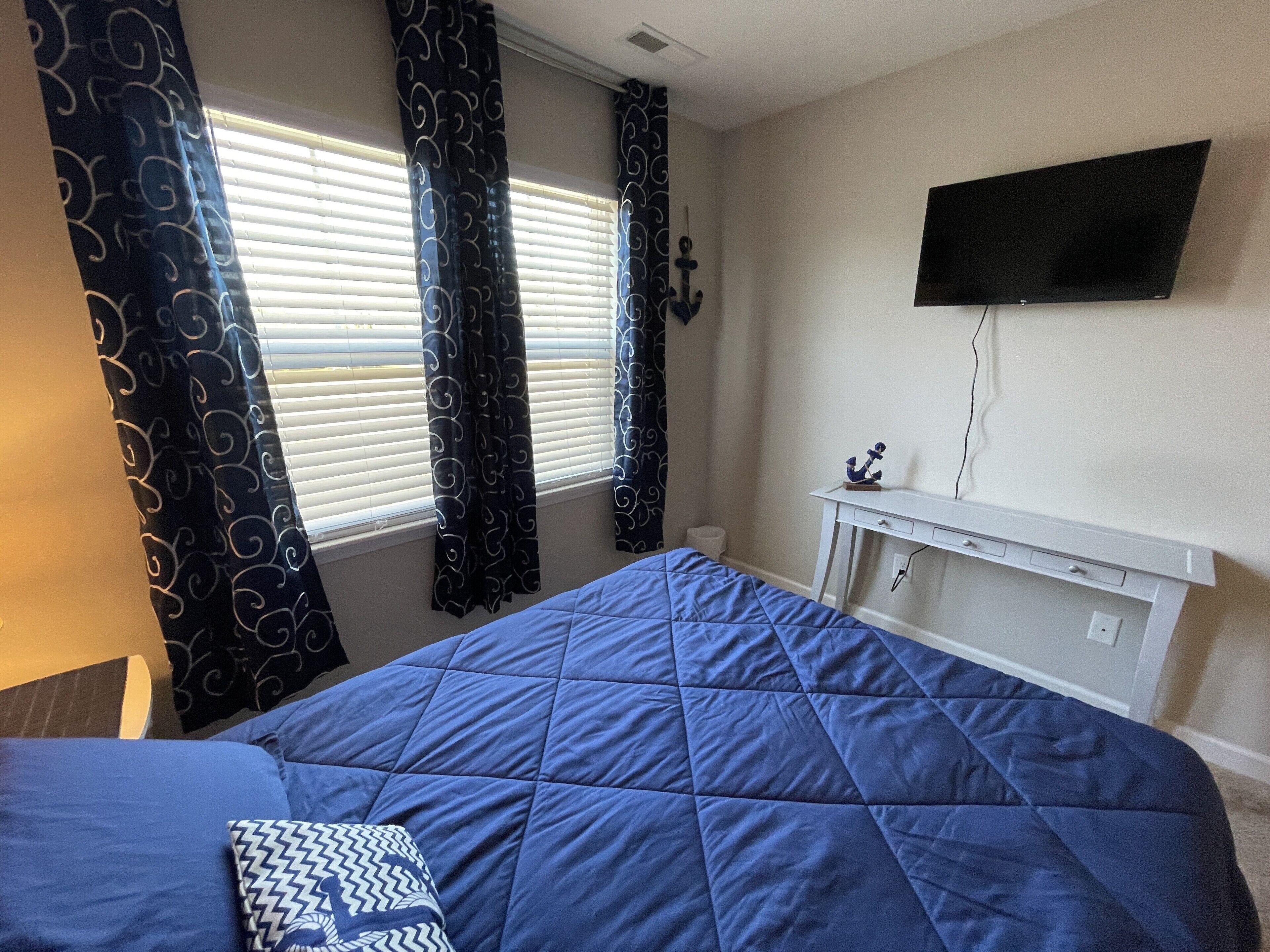 Une chambre à coucher avec un lit, un téléviseur fixé au mur, un bureau avec une chaise et des rideaux.
