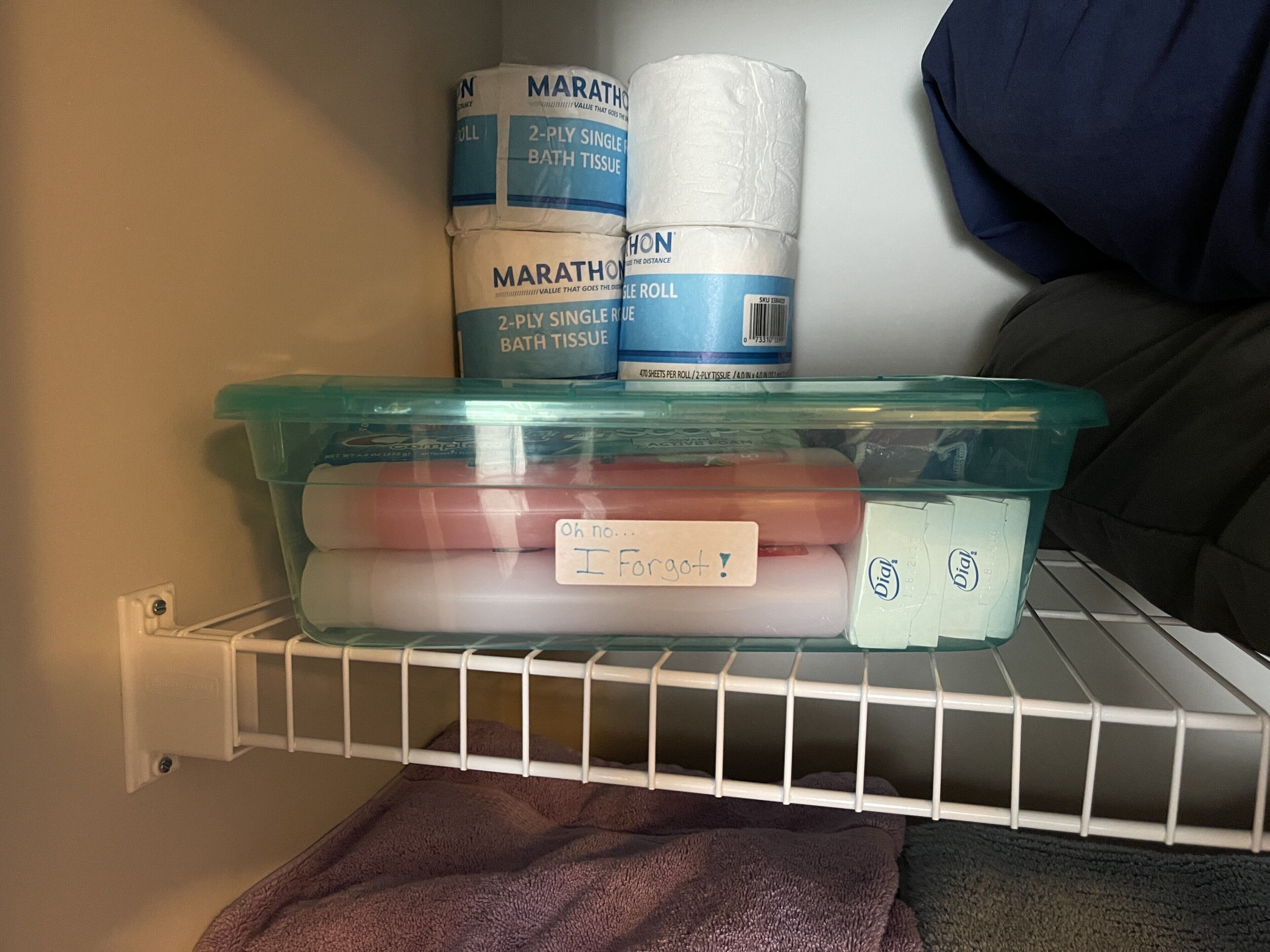 A storage shelf with a plastic container holding items labeled 'Oh no... I forgot!' and a blue container with 'MARATHON' bath tissue.