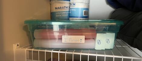 A storage shelf with a plastic container holding items labeled 'Oh no... I forgot!' and a blue container with 'MARATHON' bath tissue.