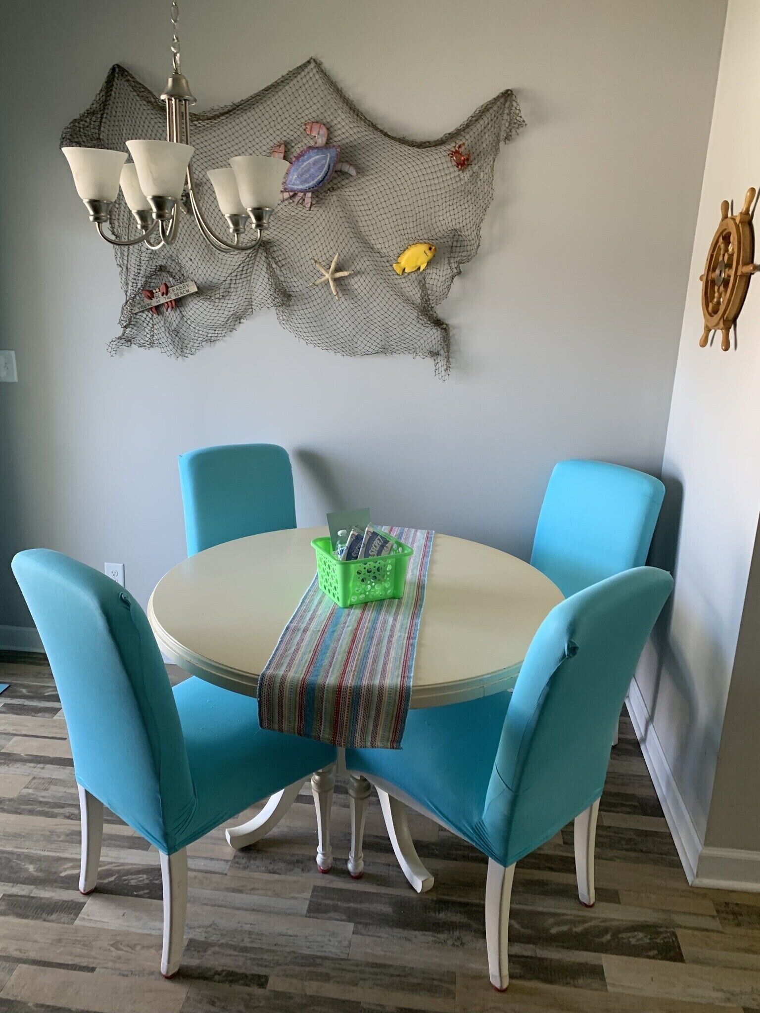 Un comedor con una mesa redonda, cuatro sillas turquesa, una cesta verde y un tapiz decorativo con motivos marinos.