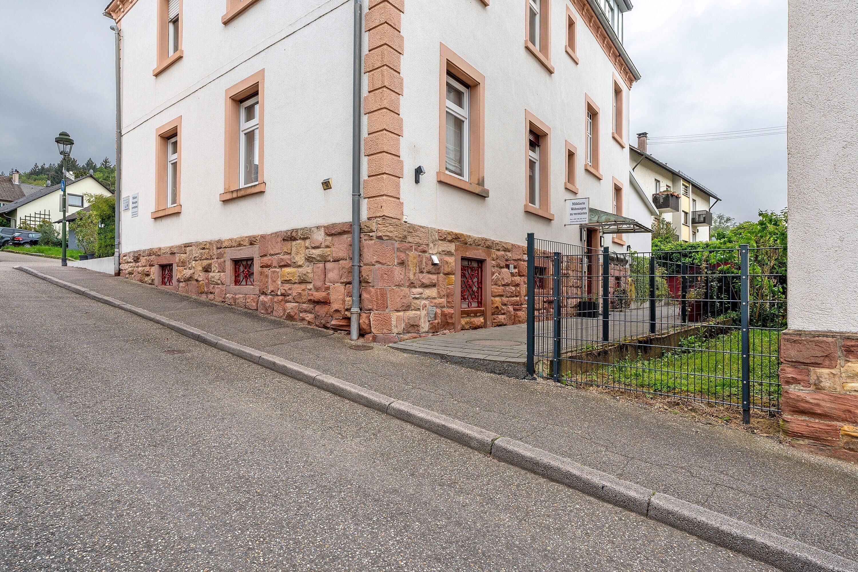 Un bâtiment dont la partie inférieure est en pierre et la partie supérieure est peinte en blanc, avec une entrée fermée par un portail et un trottoir dallé.