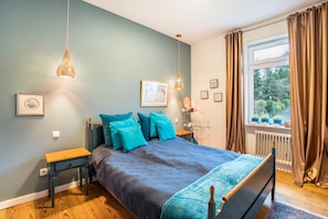 Une chambre à coucher avec un lit, des tables de chevet, une fenêtre avec des rideaux et une vue sur la verdure à l’extérieur.
