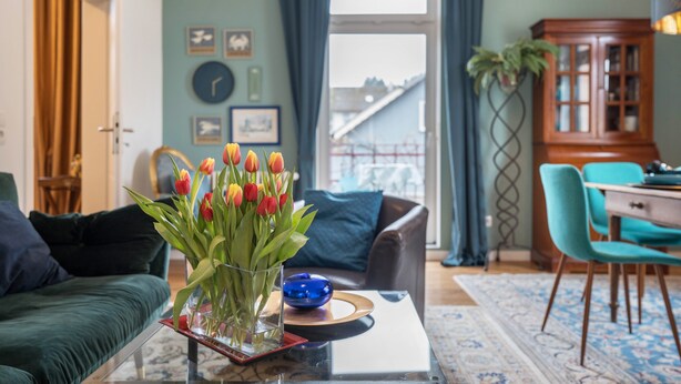 Una sala de estar con un sofá, una mesa de centro con un florero de tulipanes, un comedor con sillas y vistas por una ventana.
