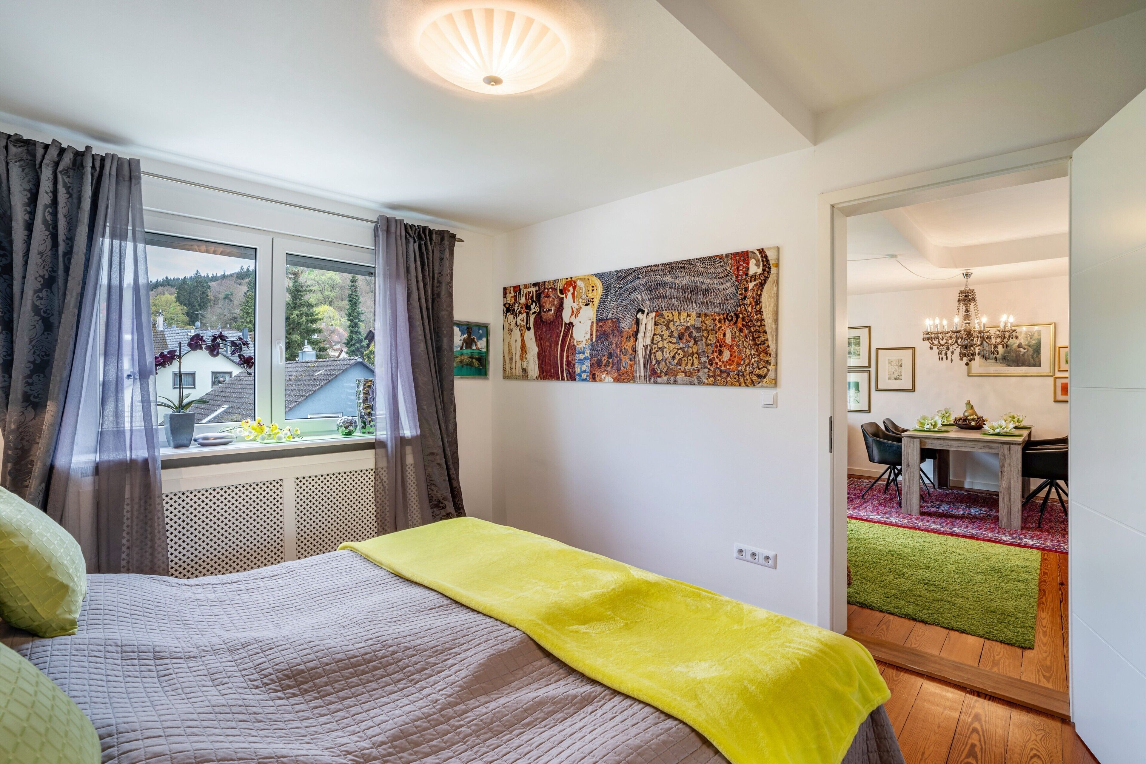 A bedroom with a bed, a window with curtains, a painting, and a dining area with a table and chairs.
