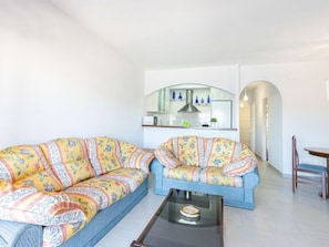 A living room with two patterned sofas, a glass coffee table, and a kitchen area with a microwave and decorative items.
