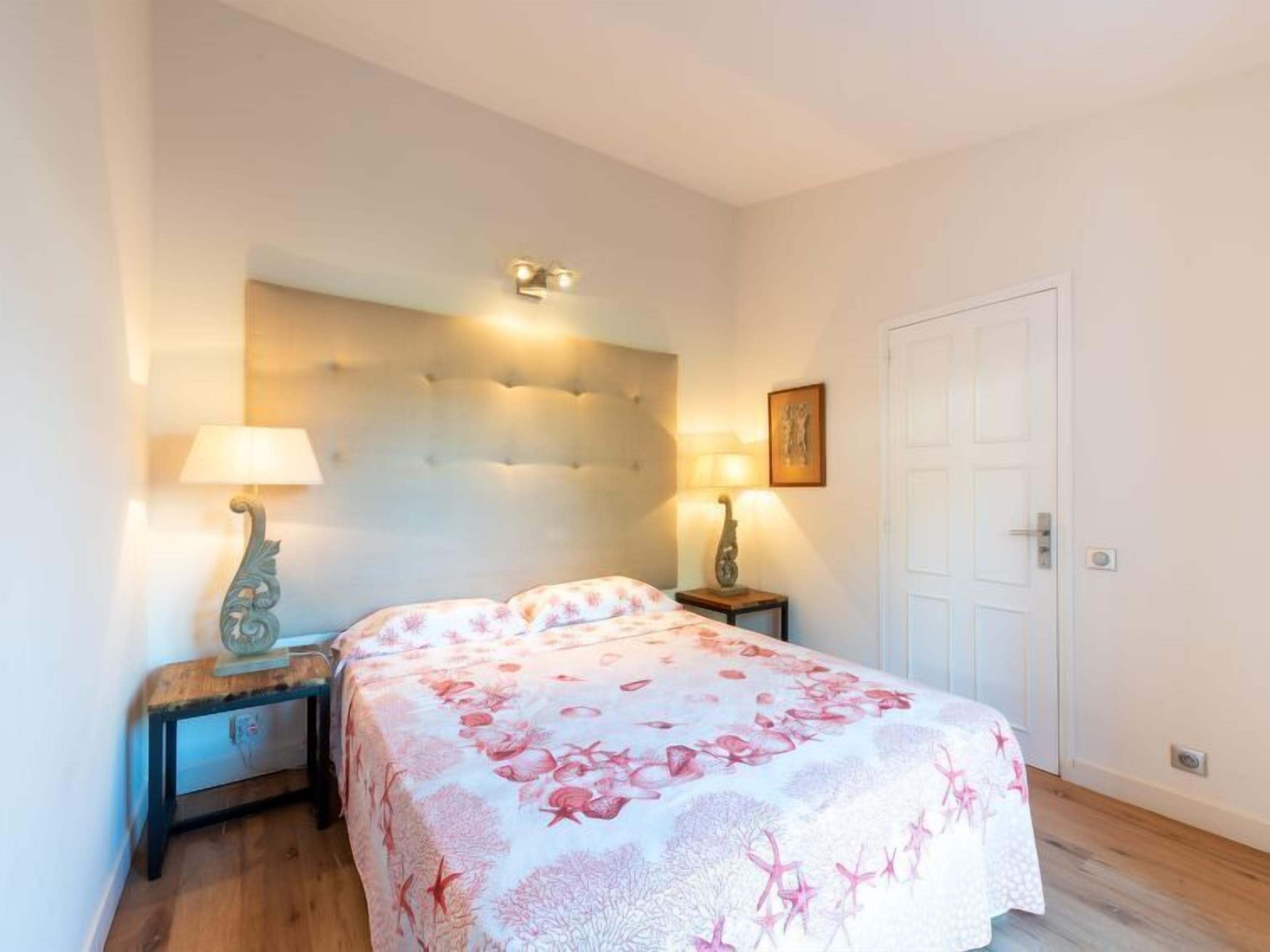 A bedroom with a tufted headboard, two bedside tables with lamps, a bed with a floral bedspread, and a white door.