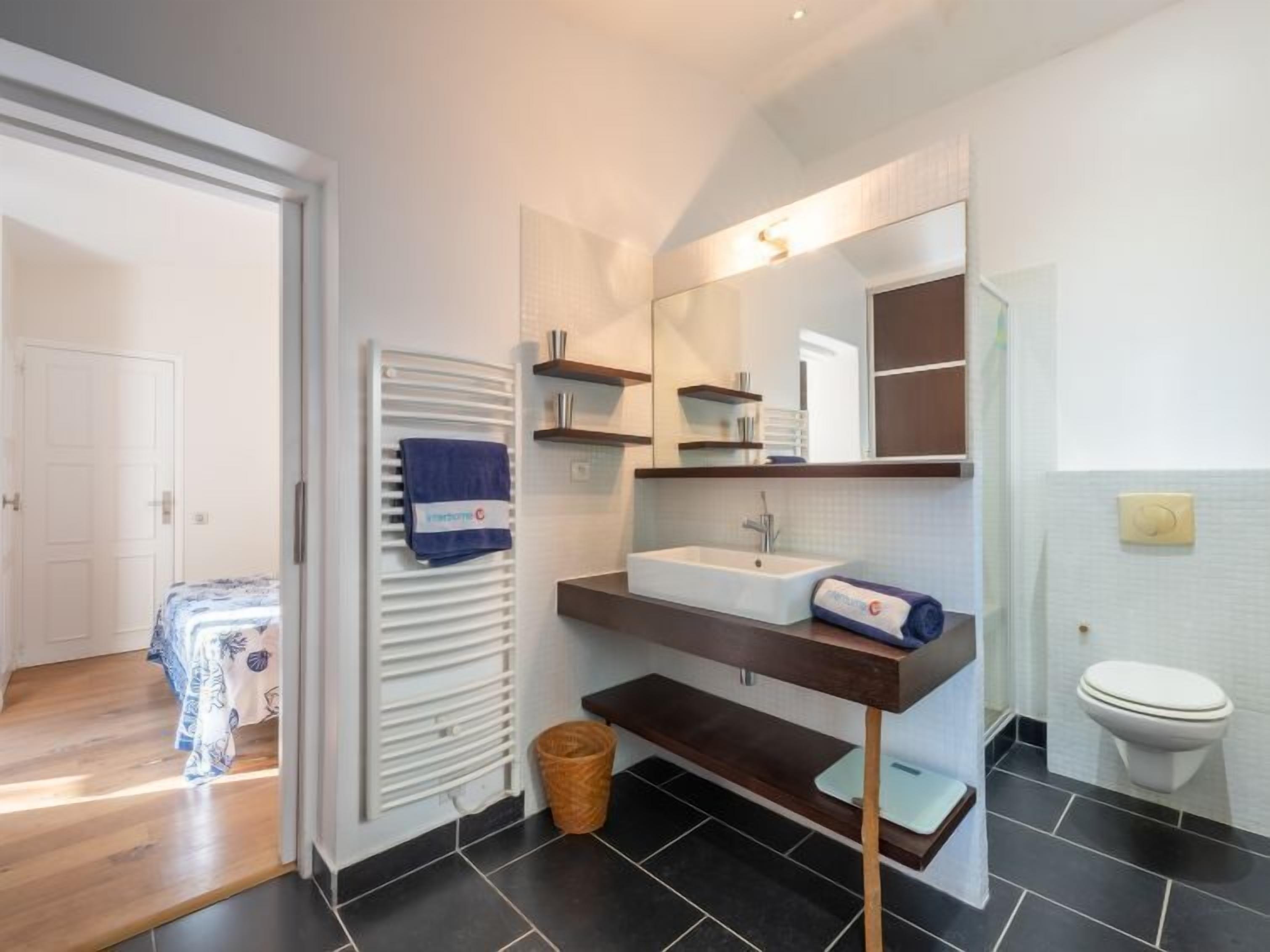 Une salle de bain moderne avec un lavabo blanc, un miroir et des toilettes. On y trouve un porte-serviettes sur lequel est suspendue une serviette bleue, ainsi qu’un petit panier posé sur le sol.