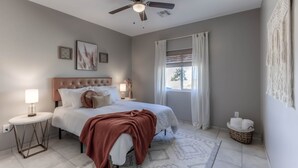 Ein Schlafzimmer mit einem Bett, einem Deckenventilator, einem Nachttisch, einem Fenster mit Vorhängen und einer Wanddekoration aus Makramee.