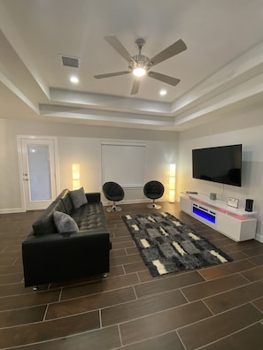 Uma sala de estar moderna com um sofá de couro preto, duas cadeiras pretas, uma grande televisão de tela plana, um ventilador de teto e um rack branco com o símbolo de Wi-Fi.