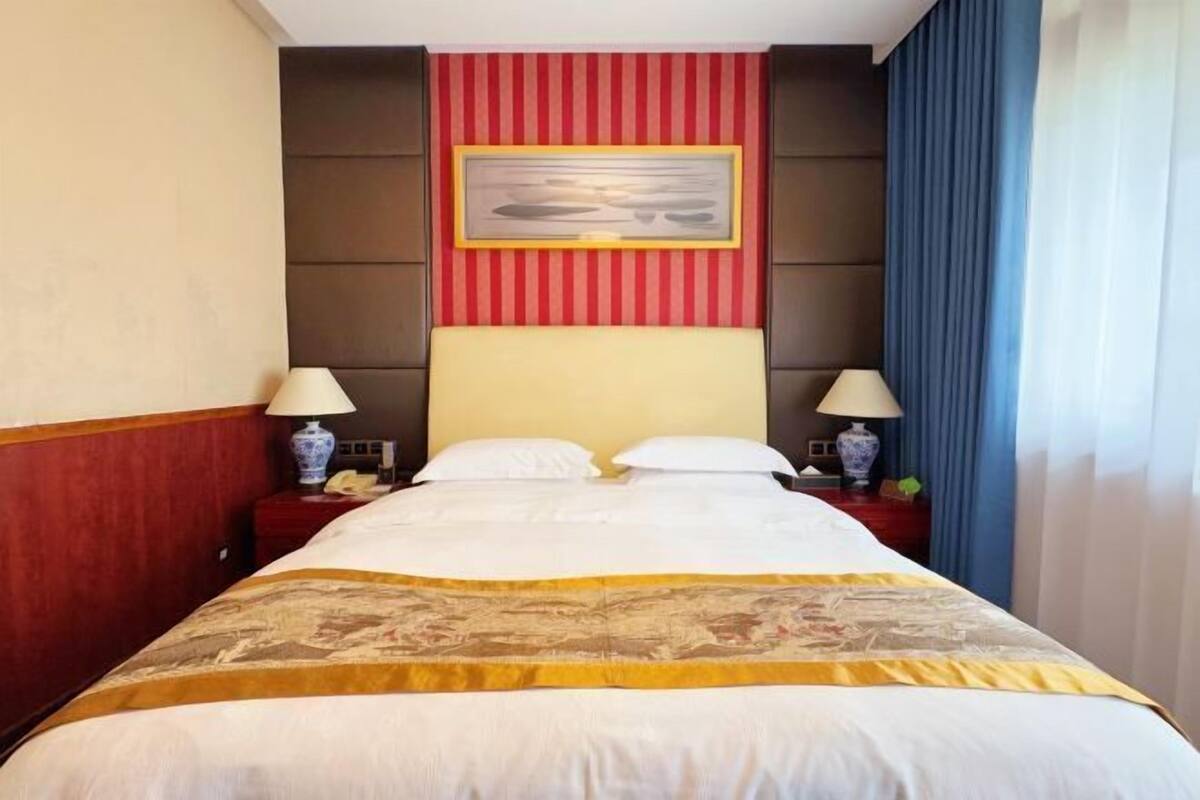 A hotel room with a bed, two bedside tables, lamps, a framed picture on the wall, and a window with curtains.