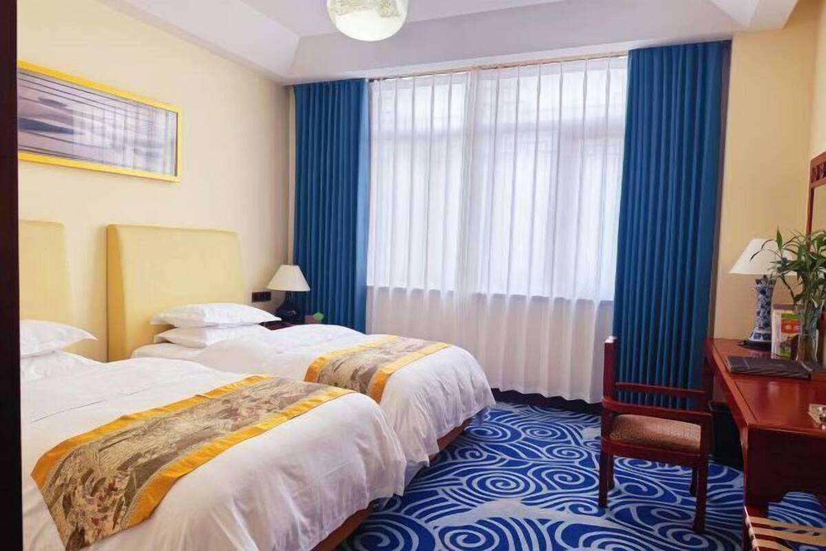 A hotel room with two beds, a desk, a chair, a lamp, and a window with blue curtains.