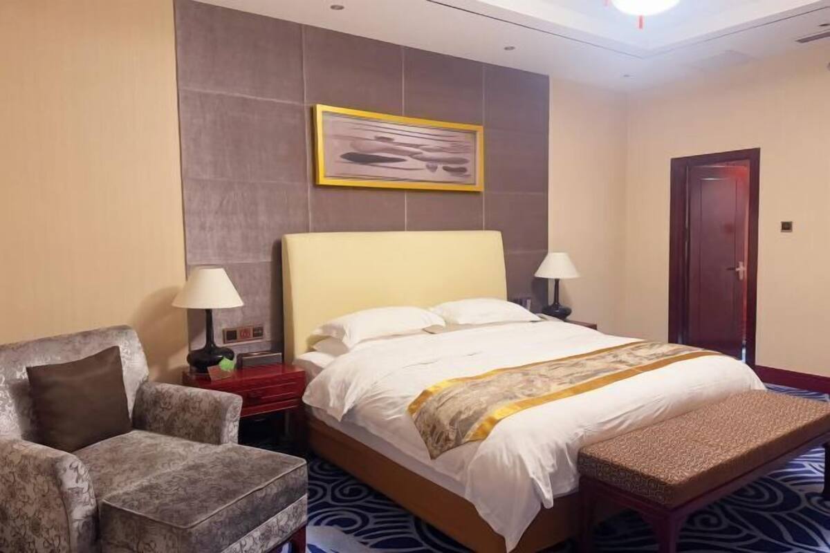 A hotel room with a bed, a chair, a bench, a nightstand, a lamp, a door, and a framed picture on the wall.