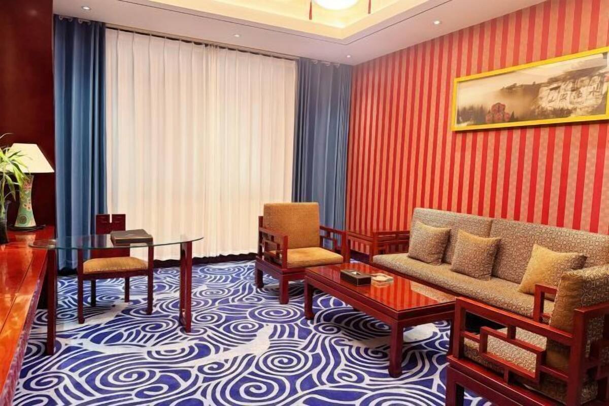 A hotel room with a patterned carpet, a sofa, two armchairs, a small table, and a painting on the wall.