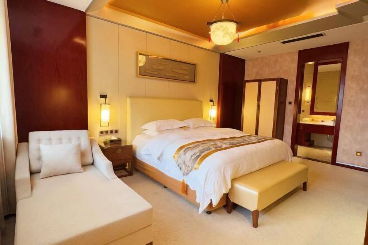 A hotel room with a large bed, a bedside table, a bench, a chair, and a mirror.