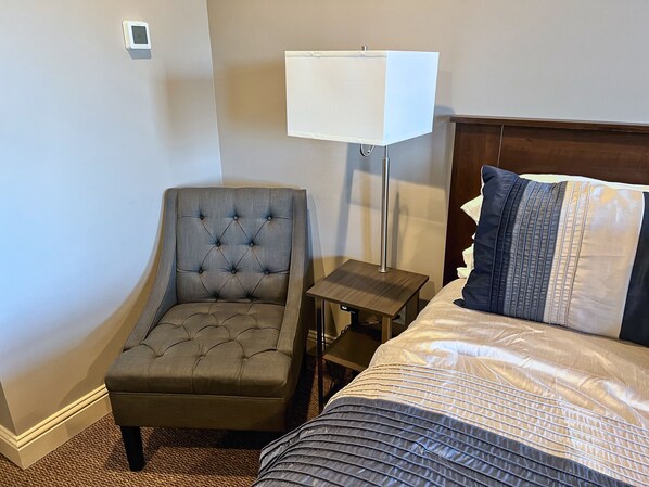 A hotel room with a tufted armchair, a bedside table with a lamp, and a bed with a patterned bedspread.