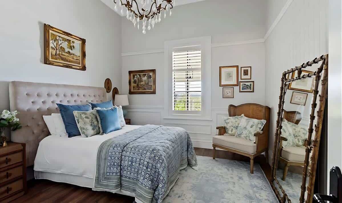 A bedroom with a tufted headboard, a bed with patterned bedding, a wooden chair, and a mirror with a wooden frame.