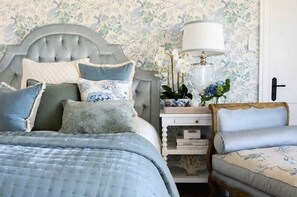 A bedroom with a tufted headboard, a bed with a quilted bedspread, a bedside table with a lamp, and a chair with floral upholstery. - Hamptons Farm Estate—Luxe Retreat & Elegant Events (Wongamine)