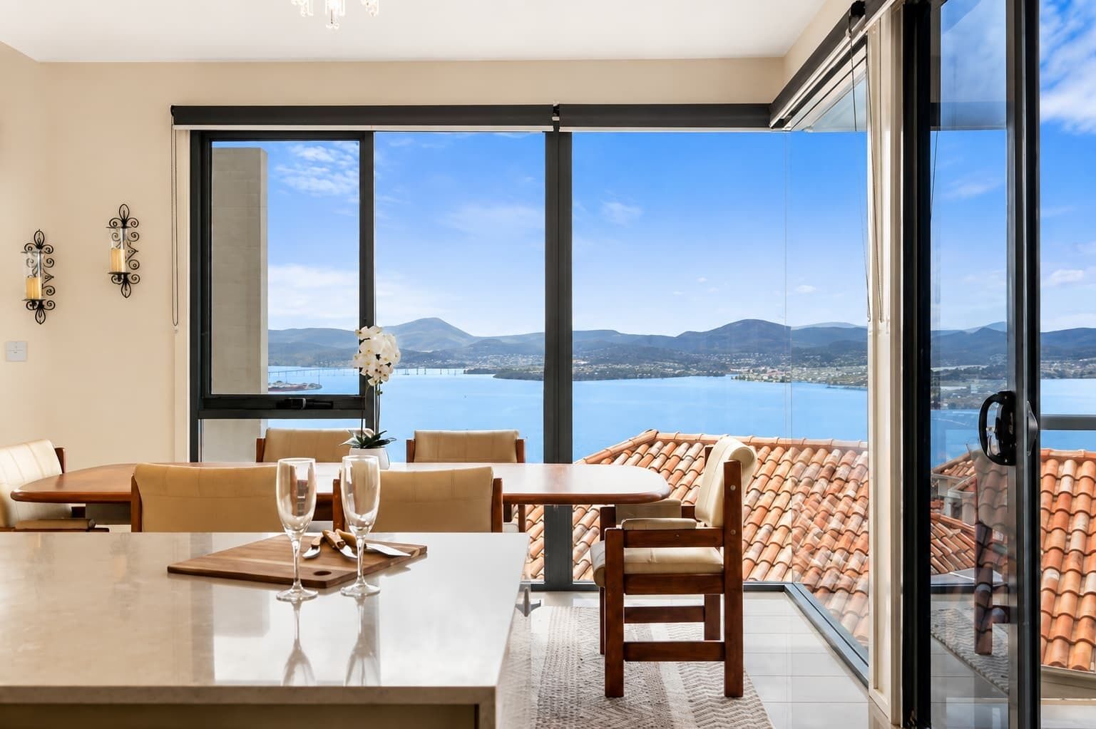 A dining area with a view of a body of water and mountains, featuring a table with chairs and a vase of flowers.