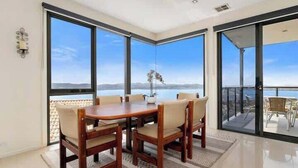 Un comedor con una mesa y sillas de madera, ventanas amplias con vistas al mar y a las montañas, y un balcón con mobiliario exterior.