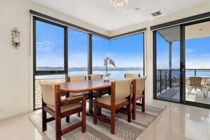 Un comedor con una mesa y sillas de madera, ventanas amplias con vistas al mar y a las montañas, y un balcón con mobiliario exterior.