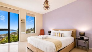 Un dormitorio con una cama grande, una mesita de noche, una silla y vistas a un paisaje costero.