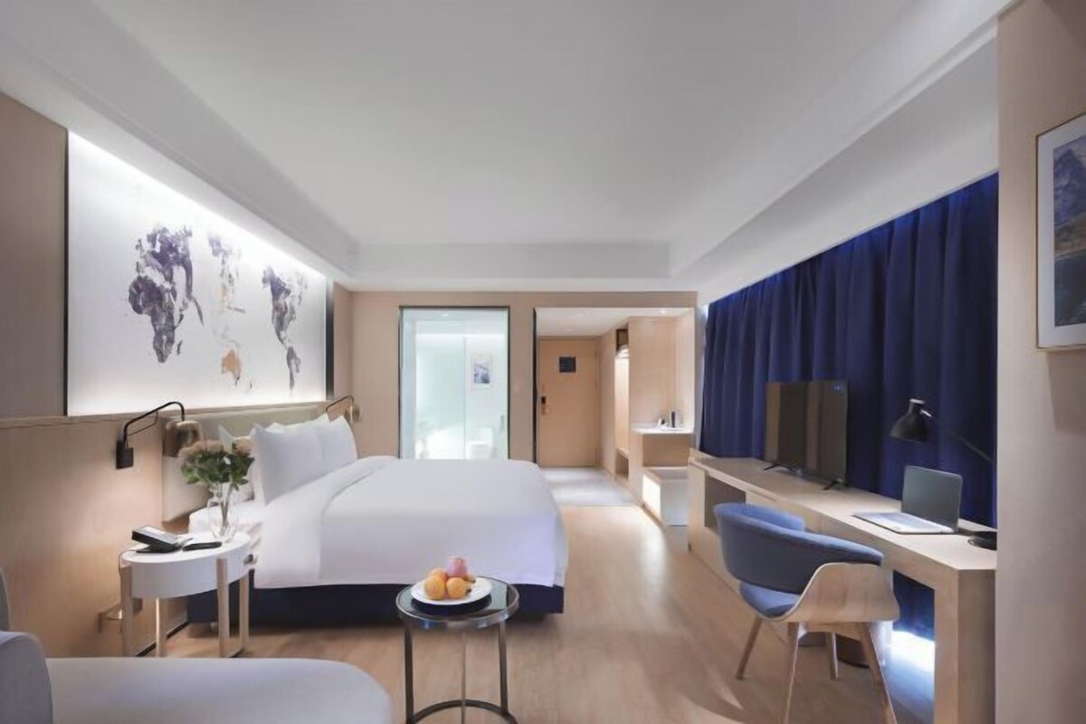 A modern hotel room with a large bed, a desk, a chair, and a small table with fruit.
