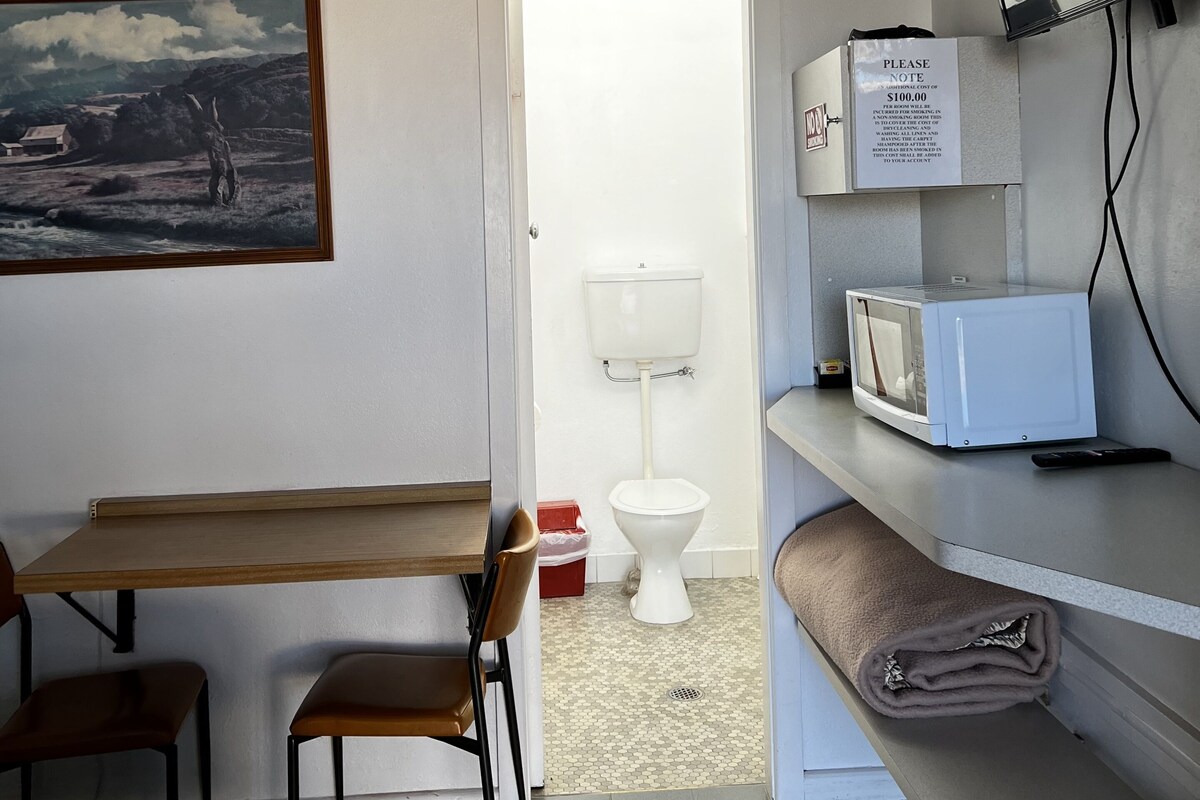 Habitación de hotel con cama, escritorio, silla, un cuadro, inodoro, microondas y televisor.