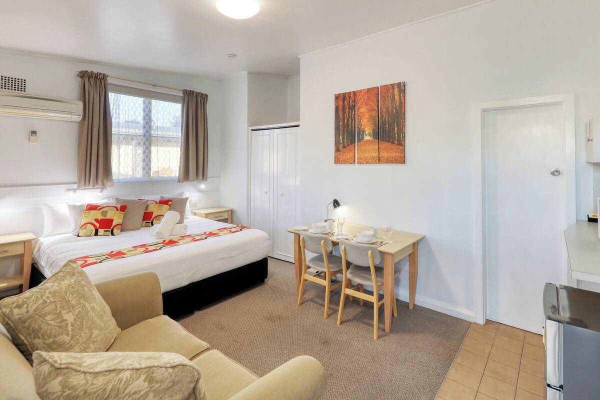 A hotel room with a bed, a dining table with chairs, a microwave, and a painting on the wall.