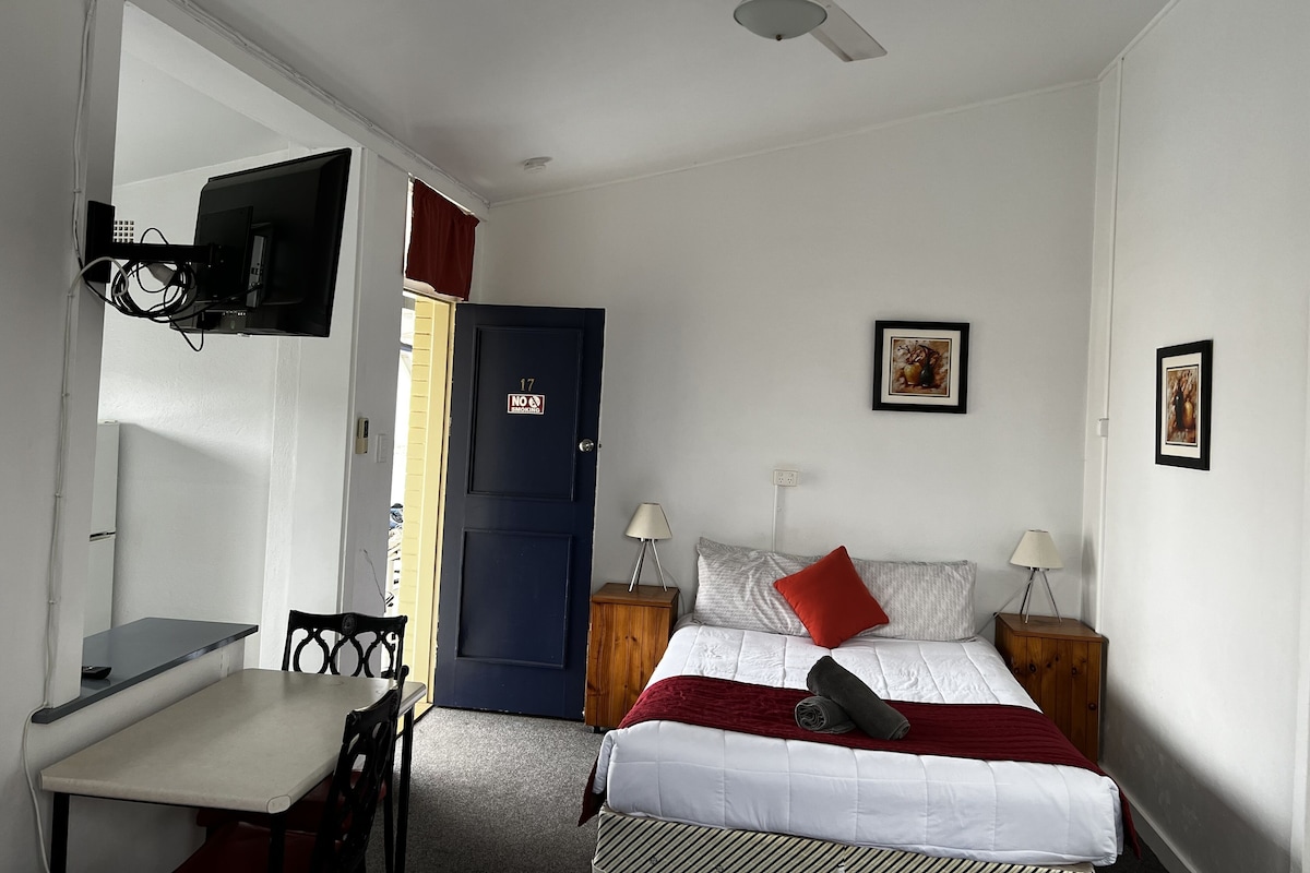 A bedroom with a bed, a desk, a chair, a TV, a ceiling fan, and a door with a 'No Smoking' sign.