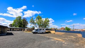 Un camping con espacio para vehículos recreativos, un edificio con zona cubierta y árboles al fondo.