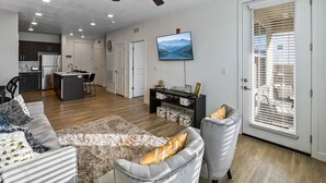 A modern living room with a grey sofa, a coffee table, and a grey armchair. There is a TV mounted on the wall, a ceiling fan, and a kitchen area in the background.