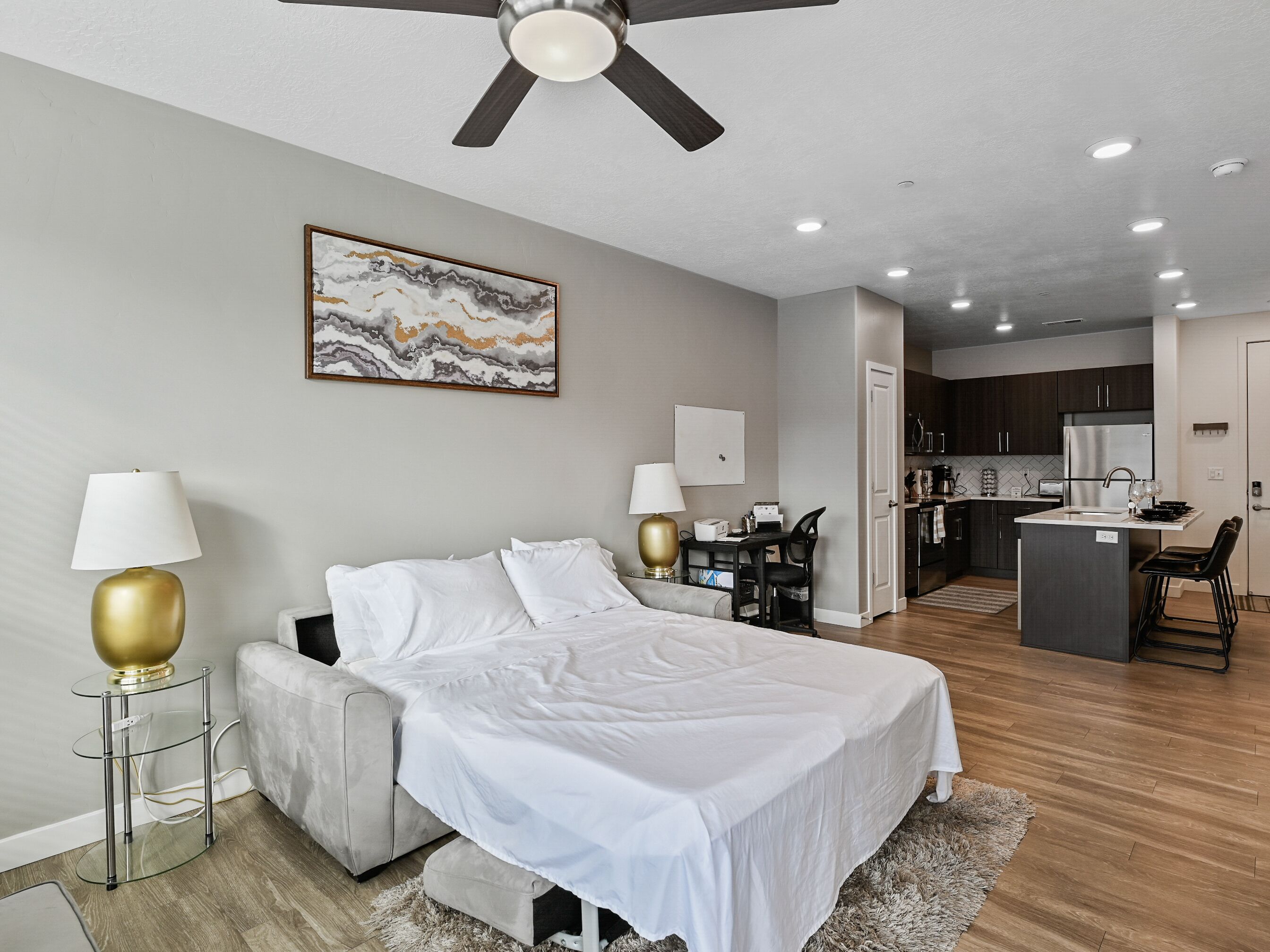 A modern hotel room with a bed, bedside lamps, a desk, and a kitchenette.