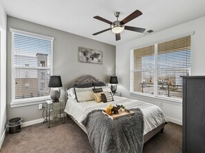 A bedroom with a bed, two bedside tables, a ceiling fan, and large windows with blinds.