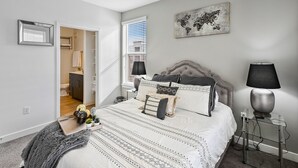 A bedroom with a bed, bedside lamps, a world map artwork, a ceiling fan, and a view of the bathroom.