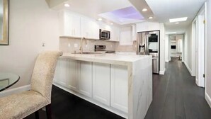 A modern kitchen with white cabinetry, a central island, and a dining area with a chair. - Gorgeous Beachfront  Condo (Imperial Beach)