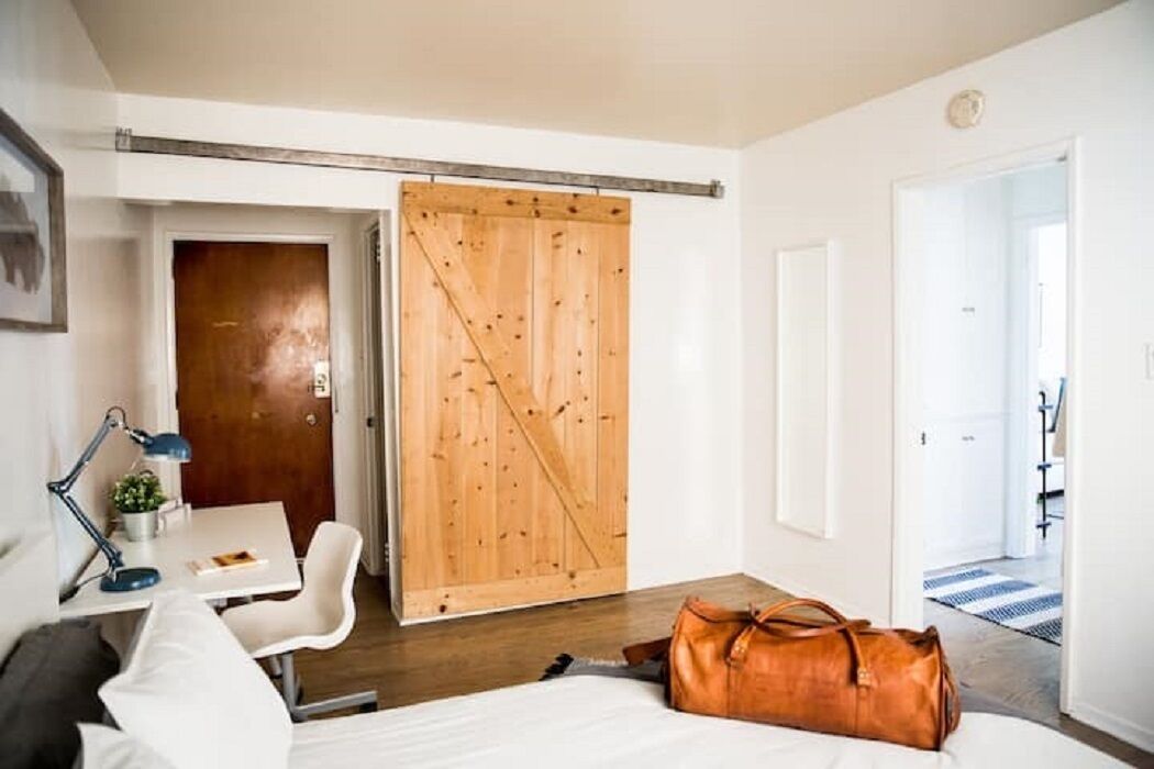 Ein Zimmer mit einem Bett, einem Schreibtisch mit Lampe, einem Stuhl, einer Holztür, die sich schiebt, und einer braunen Tasche auf dem Boden.
