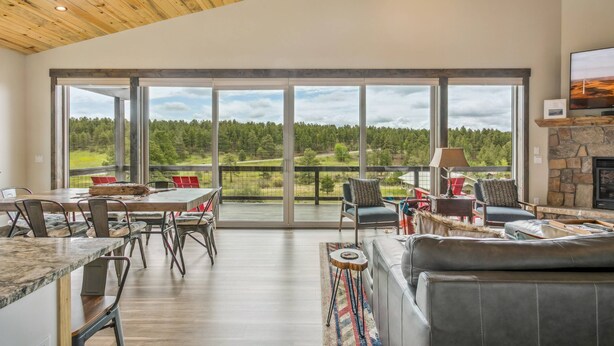 A spacious living area with a large window overlooking a forested landscape, a stone fireplace, and a modern dining setup.