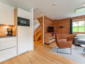 A modern kitchen with a built-in microwave, wooden flooring, a staircase leading to a loft, a grey sofa, and a large window with a view of greenery.