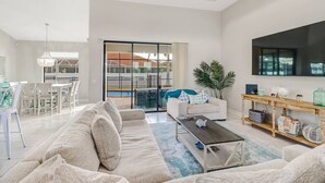 Un salón espacioso con un sofá grande, una mesa de centro y un televisor montado en la pared. Hay una puerta corrediza de vidrio que da a un balcón con vista a la piscina.