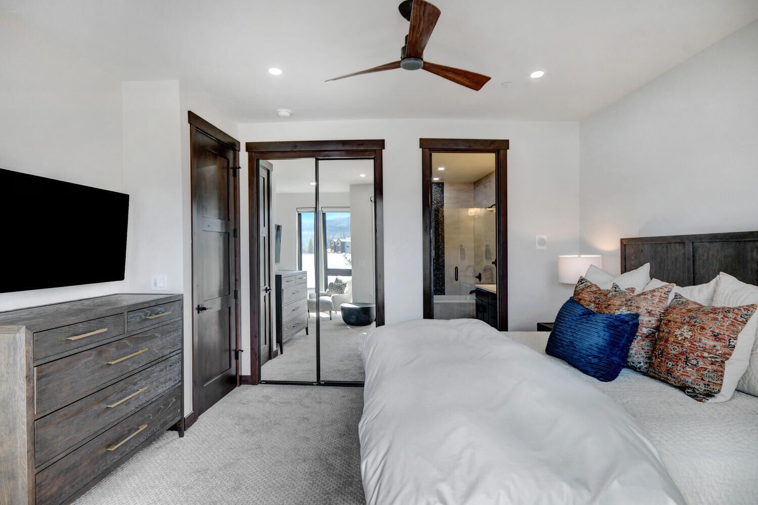 A bedroom with a large bed, a TV mounted on the wall, a ceiling fan, and a view of the outside through a glass door.
