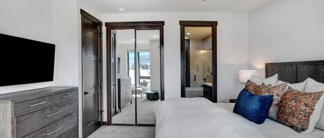 A bedroom with a large bed, a TV mounted on the wall, a ceiling fan, and a view of the outside through a glass door.