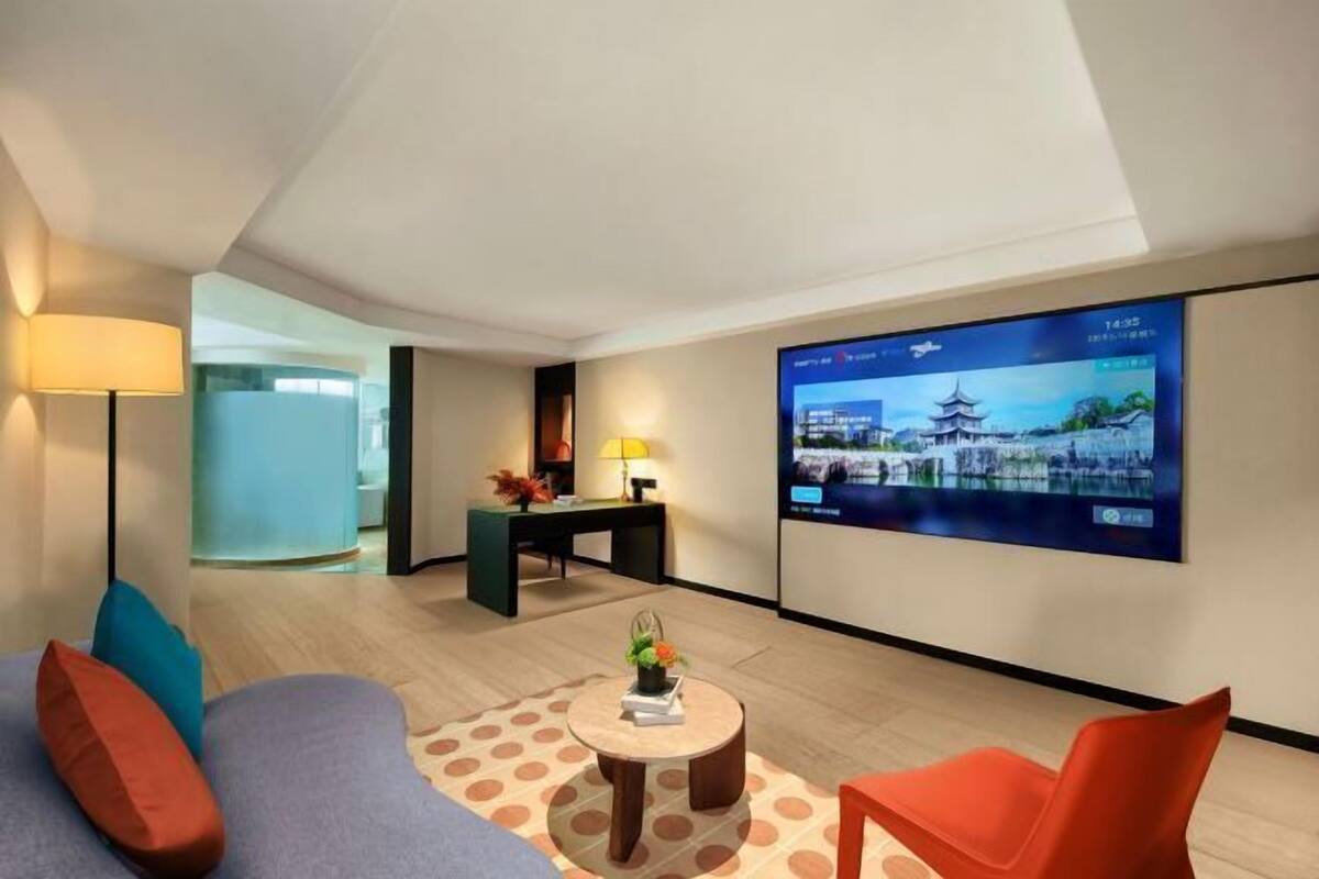 A modern hotel room with a large flat-screen TV displaying a scenic view, a comfortable sofa, a small round coffee table, and a desk with a lamp.
