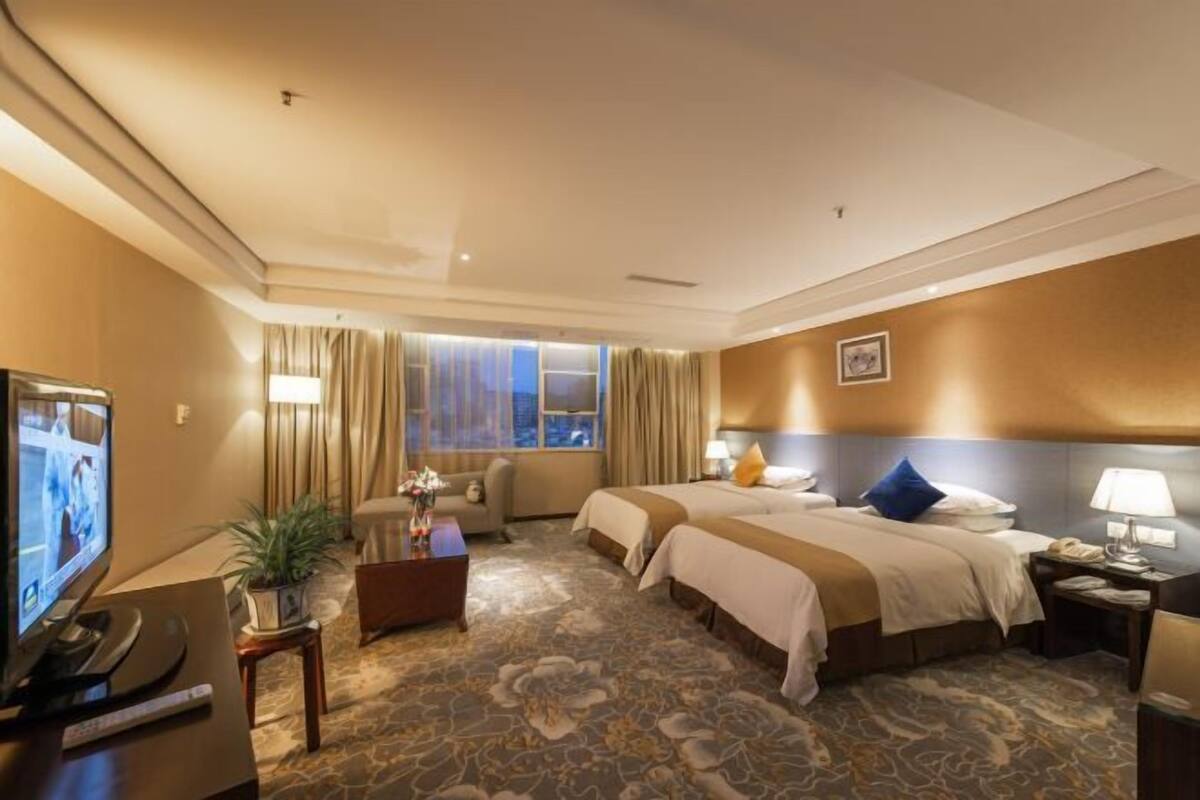 A hotel room with two beds, a TV, a desk with a lamp, and a window with curtains.
