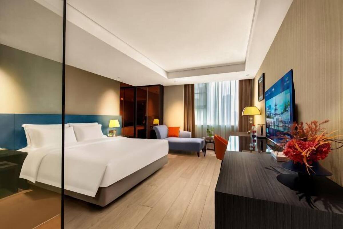 A modern hotel room with a large bed, a desk, a chair, a TV, and a window with curtains.