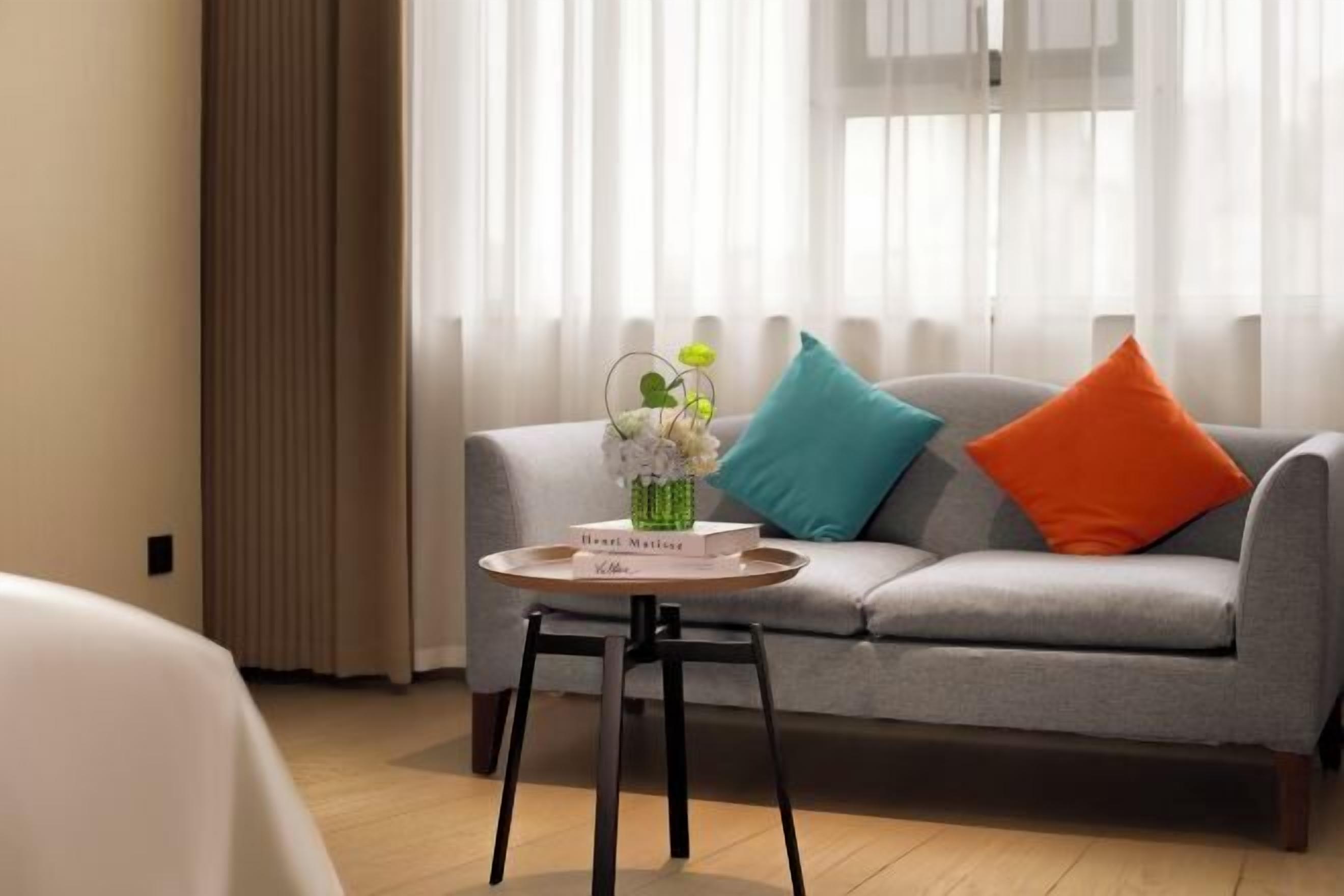Une chambre d’hôtel moderne avec un canapé gris, des coussins turquoise et orange, une petite table ronde avec un plateau, et un vase contenant des fleurs blanches.