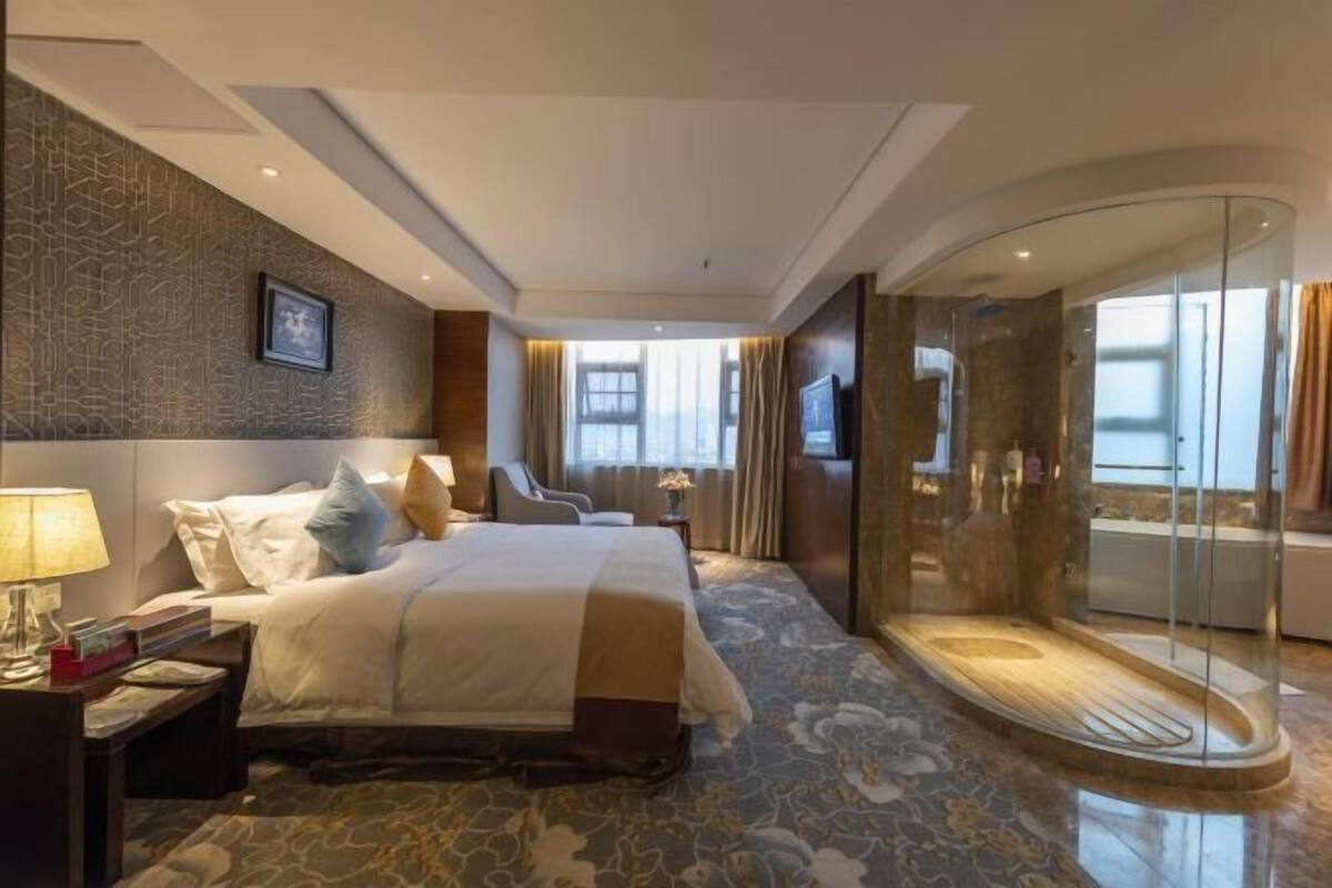 A hotel room with a large bed, a glass-enclosed shower, a desk with a lamp, and a view of the city.