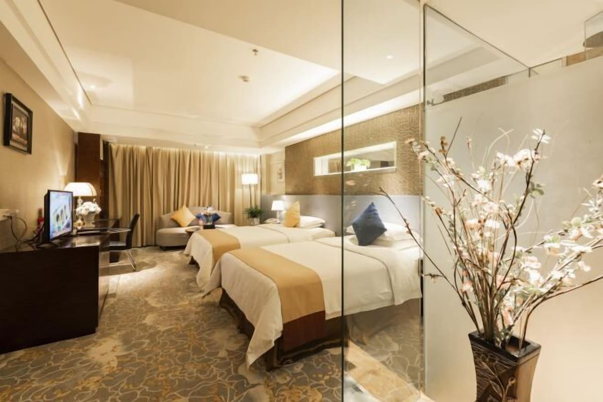 A hotel room with two beds, a desk, a chair, a glass partition, and a vase with flowers.